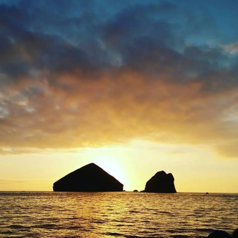 Sunset over ocean with two large silhouetted rocks on the horizon and cloudy sky.
