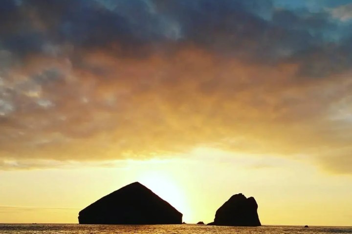 Sunset over ocean with two large silhouetted rocks on the horizon and cloudy sky.