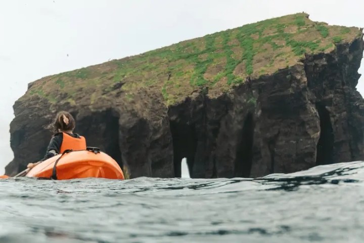 Person in orange kayak near large rocky island with grass patches.