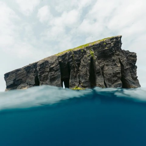 Rocky island with vertical cliffs rises above blue ocean water under cloudy sky.