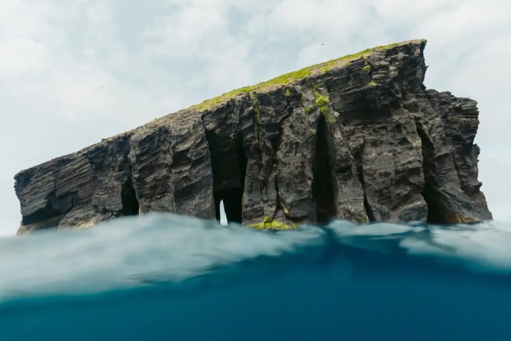 Rocky island with vertical cliffs rises above blue ocean water under cloudy sky.