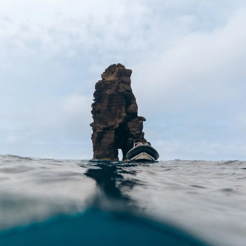 Rock formation in ocean with small boat nearby under cloudy sky.