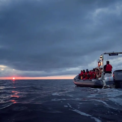 Boat with people on ocean at sunset under cloudy sky.