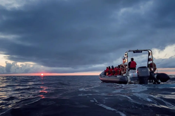 Boat with people on ocean at sunset under cloudy sky.
