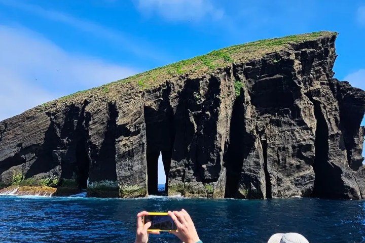 Tourists on a boat photograph a rocky island with sea caves under a blue sky.