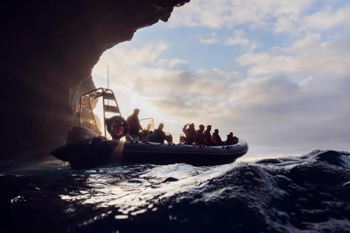 Boat with people near rocky cave entrance at sea with sunlight and waves.
